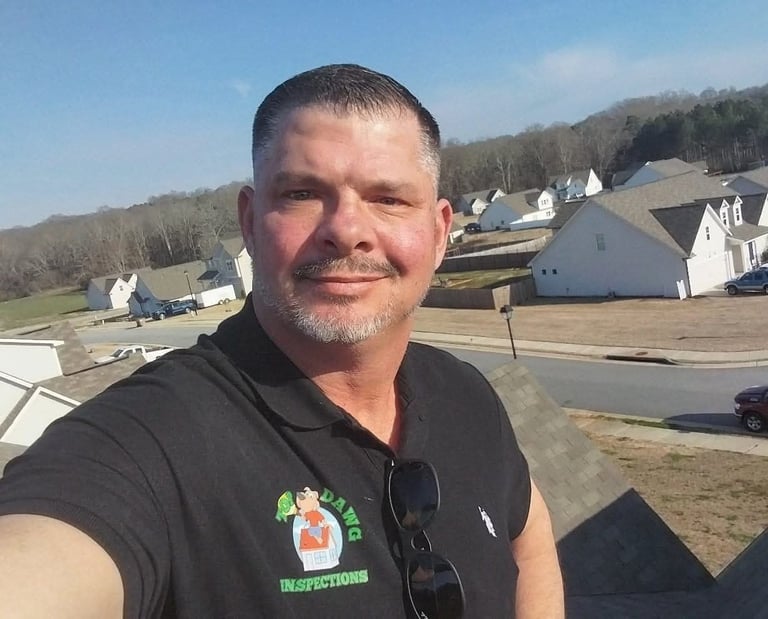 Man with short dark hair and goatee wearing black shirt taking selfie in residential neighborhood with white houses and bare trees